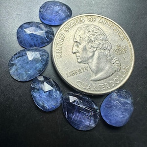 May include: Six faceted, oval-shaped blue gemstones arranged next to a United States quarter dollar coin. The stones exhibit varying shades of translucent blue with visible internal inclusions and geometric facets across their surfaces.