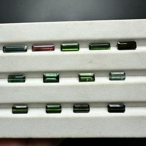 May include: A collection of green, black, and pink gemstones in a rectangular shape, displayed on a white tray.