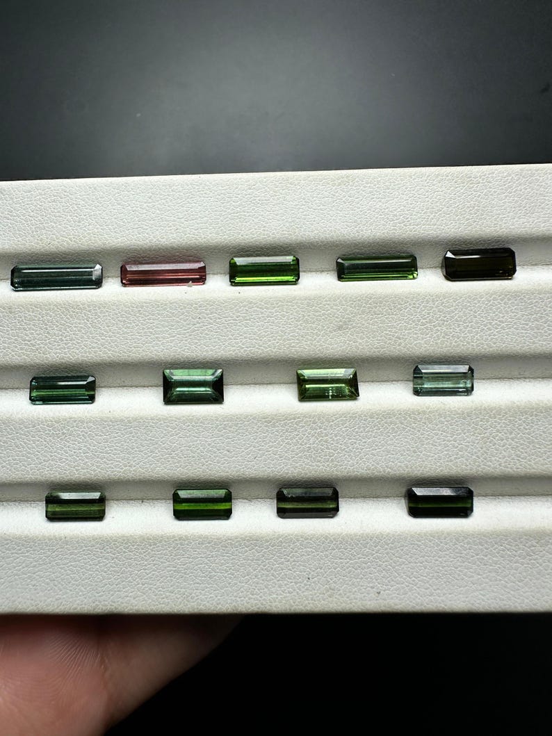 May include: A collection of green, black, and pink gemstones in a rectangular shape, displayed on a white tray.