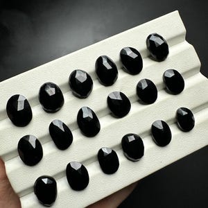 May include: A collection of 18 black oval-shaped gemstones displayed on a white tray. The gemstones have a faceted cut and a glossy finish.
