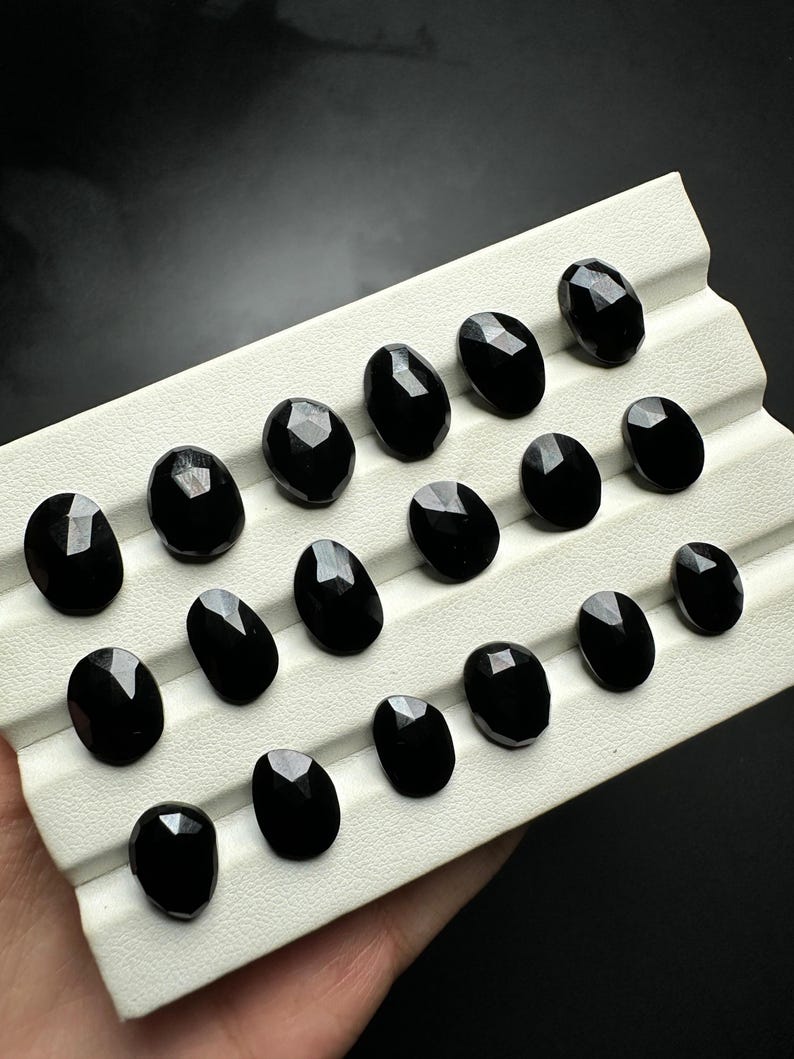 May include: A collection of 18 black oval-shaped gemstones displayed on a white tray. The gemstones have a faceted cut and a glossy finish.
