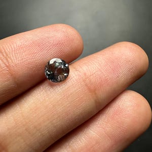 May include: A round, faceted gemstone with a dark grey colour and a hint of purple.