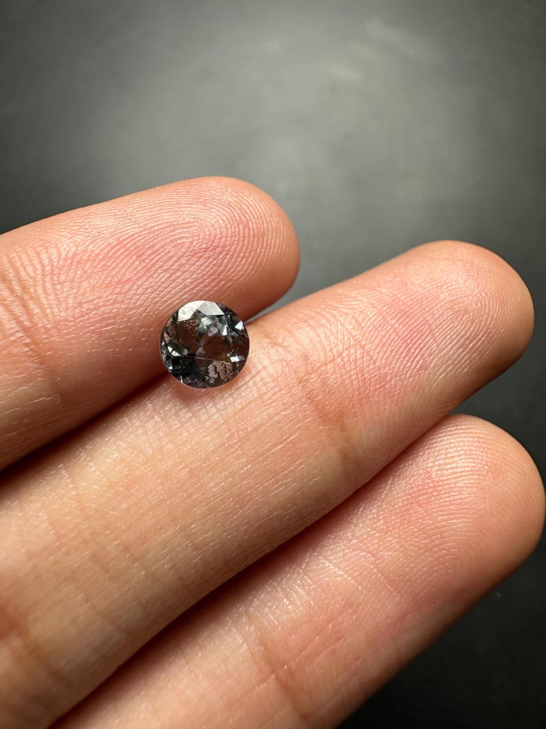 May include: A round, faceted gemstone with a dark grey colour and a hint of purple.