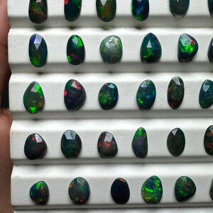 May include: A collection of 25 black opal gemstones, each with a unique green and red iridescent sheen. The opals are displayed in a white tray with individual compartments.