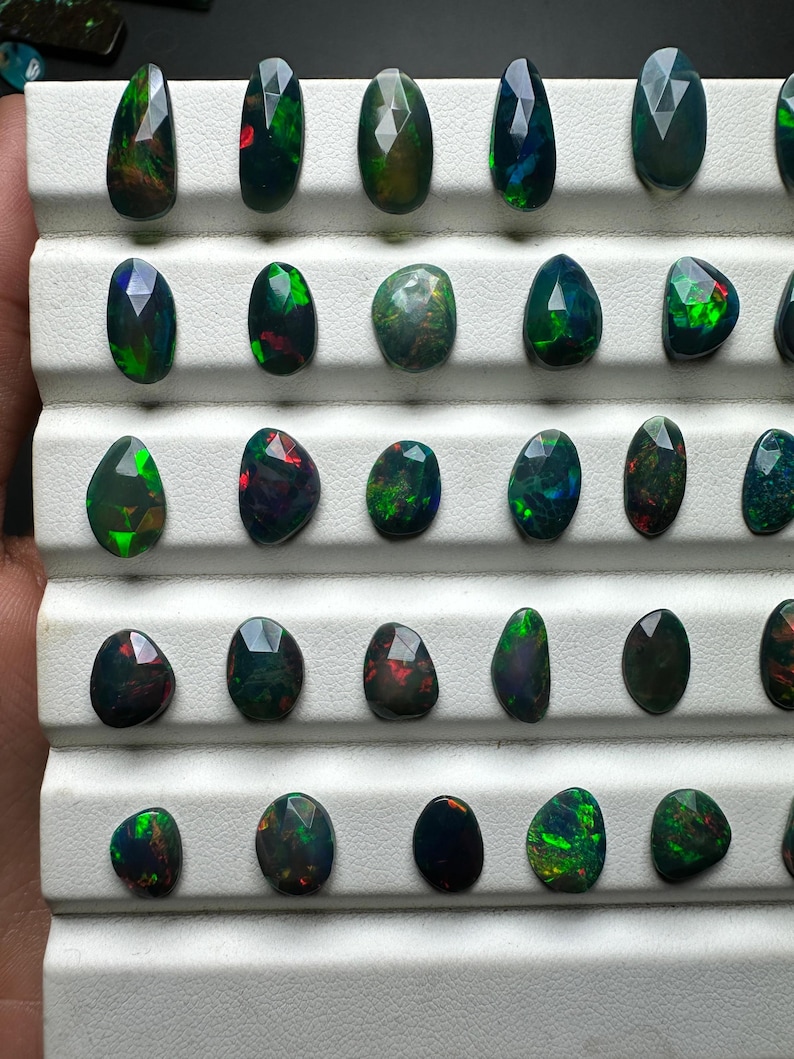 May include: A collection of 25 black opal gemstones, each with a unique green and red iridescent sheen. The opals are displayed in a white tray with individual compartments.
