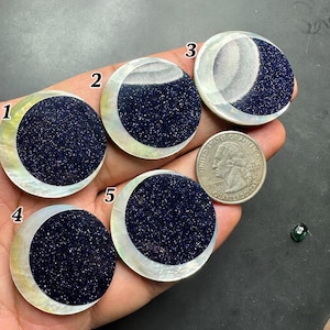 May include: Five crescent moon-shaped cabochons with a dark blue, glittery surface, set in a pearlescent, white shell. A US quarter coin is next to the cabochons. The text "1 inch (25mm)" is at the bottom.