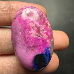 May include: An oval-shaped polished gemstone featuring a vibrant magenta and pink surface with mottled white inclusions, a distinct deep blue patch, and a small solid black circular area near the bottom edge, showcasing a smooth, glossy finish.