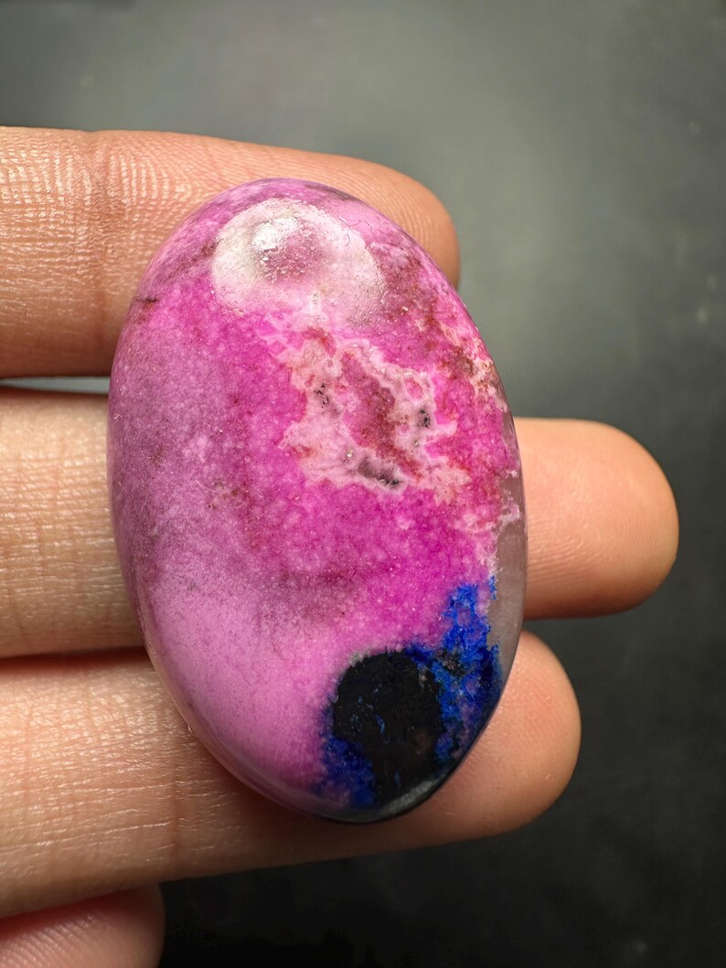 May include: An oval-shaped polished gemstone featuring a vibrant magenta and pink surface with mottled white inclusions, a distinct deep blue patch, and a small solid black circular area near the bottom edge, showcasing a smooth, glossy finish.