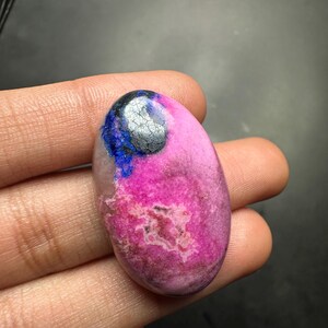 May include: An oval-shaped cabochon gemstone featuring a vibrant magenta pink base with a distinct patch of deep blue and a metallic silver-grey inclusion at the top. The stone has a smooth, polished surface with natural mineral textures.