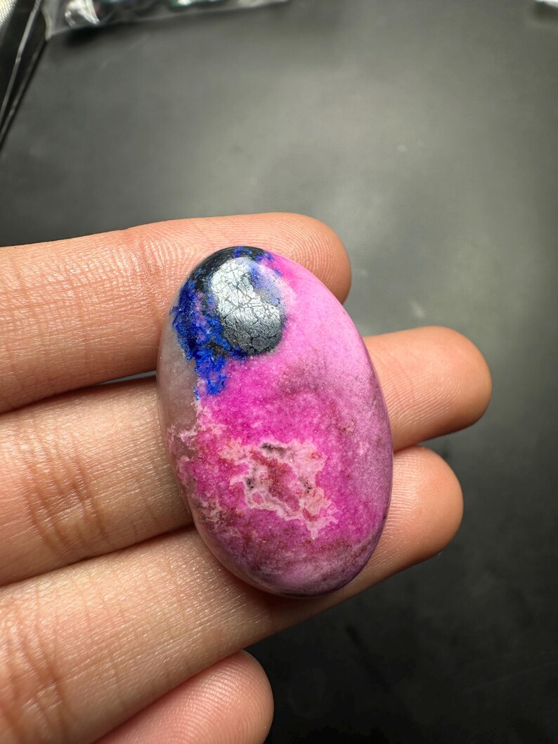 May include: An oval-shaped cabochon gemstone featuring a vibrant magenta pink base with a distinct patch of deep blue and a metallic silver-grey inclusion at the top. The stone has a smooth, polished surface with natural mineral textures.