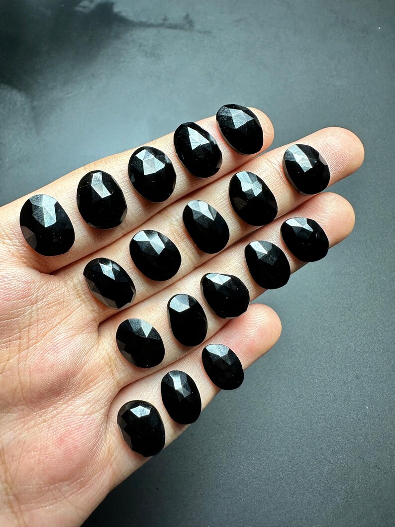 May include: A collection of 15 black faceted oval gemstones. The stones are arranged in rows on a hand.