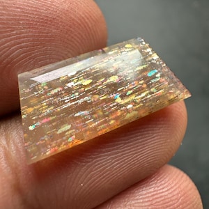 Op de afbeelding: Een rechthoekige, doorschijnende zonnesteen edelsteen met een gepolijst, plat oppervlak. De steen vertoont interne insluitingen die een glinsterend, veelkleurig effect creëren met vlekjes van goud, oranje, roze en blauw licht door het lichaam.