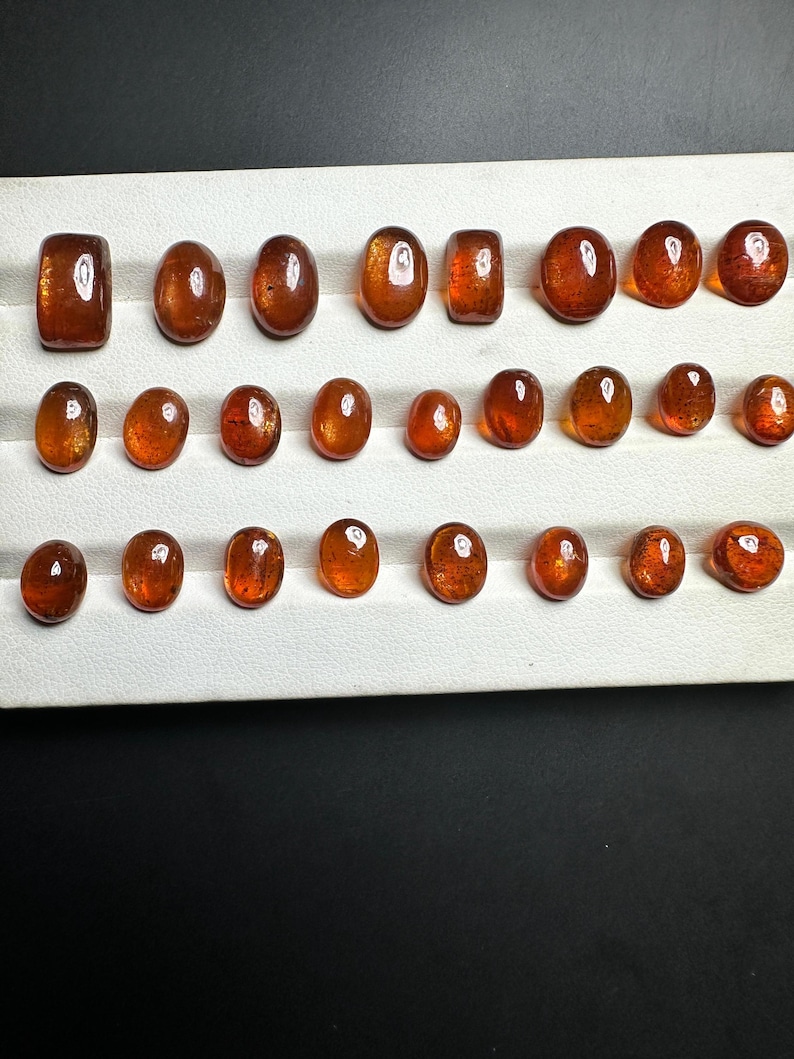 May include: A collection of 24 oval-shaped gemstones, ranging in size and color, displayed on a white tray. The gemstones are a deep orange-brown color with a slight shimmer.