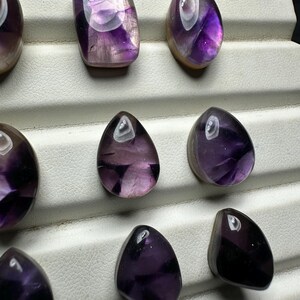 May include: An assortment of polished amethyst gemstones in various shapes, including oval and teardrop. The stones display a range of purple hues with translucent and opaque sections. The gemstones are arranged on a white display surface.