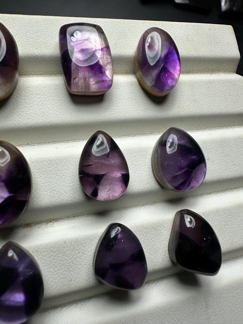 May include: An assortment of polished amethyst gemstones in various shapes, including oval and teardrop. The stones display a range of purple hues with translucent and opaque sections. The gemstones are arranged on a white display surface.