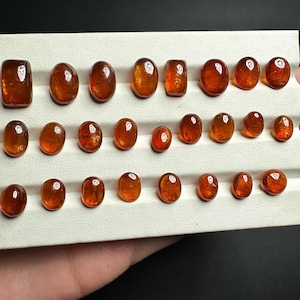 May include: A collection of 25 oval-shaped, translucent, orange-brown gemstones displayed on a white tray. The gemstones are arranged in rows and are of varying sizes.