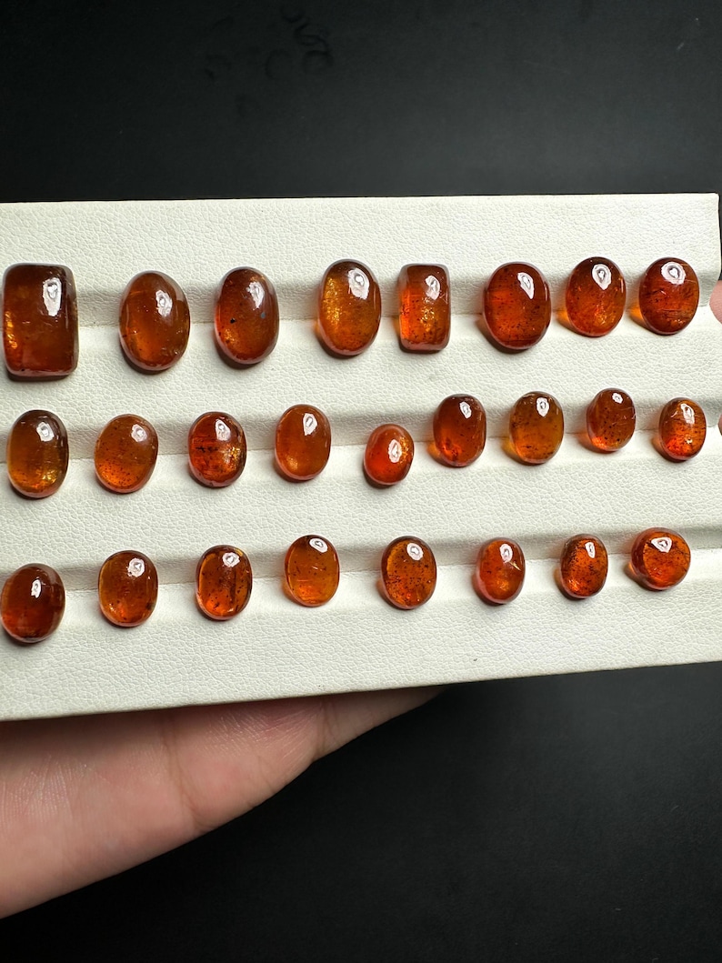 May include: A collection of 25 oval-shaped, translucent, orange-brown gemstones displayed on a white tray. The gemstones are arranged in rows and are of varying sizes.