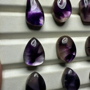 May include: A collection of polished amethyst gemstones in various shapes, including teardrop and oval. The stones display a range of purple hues, from deep violet to lighter shades, with a glossy finish. The gemstones are arranged on a white surface.