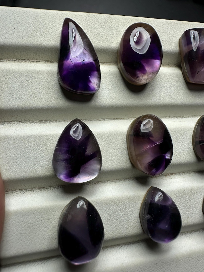 May include: A collection of polished amethyst gemstones in various shapes, including teardrop and oval. The stones display a range of purple hues, from deep violet to lighter shades, with a glossy finish. The gemstones are arranged on a white surface.