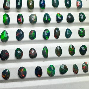 May include: A collection of 36 black opals with green and red flashes. The opals are displayed in rows on a white surface.