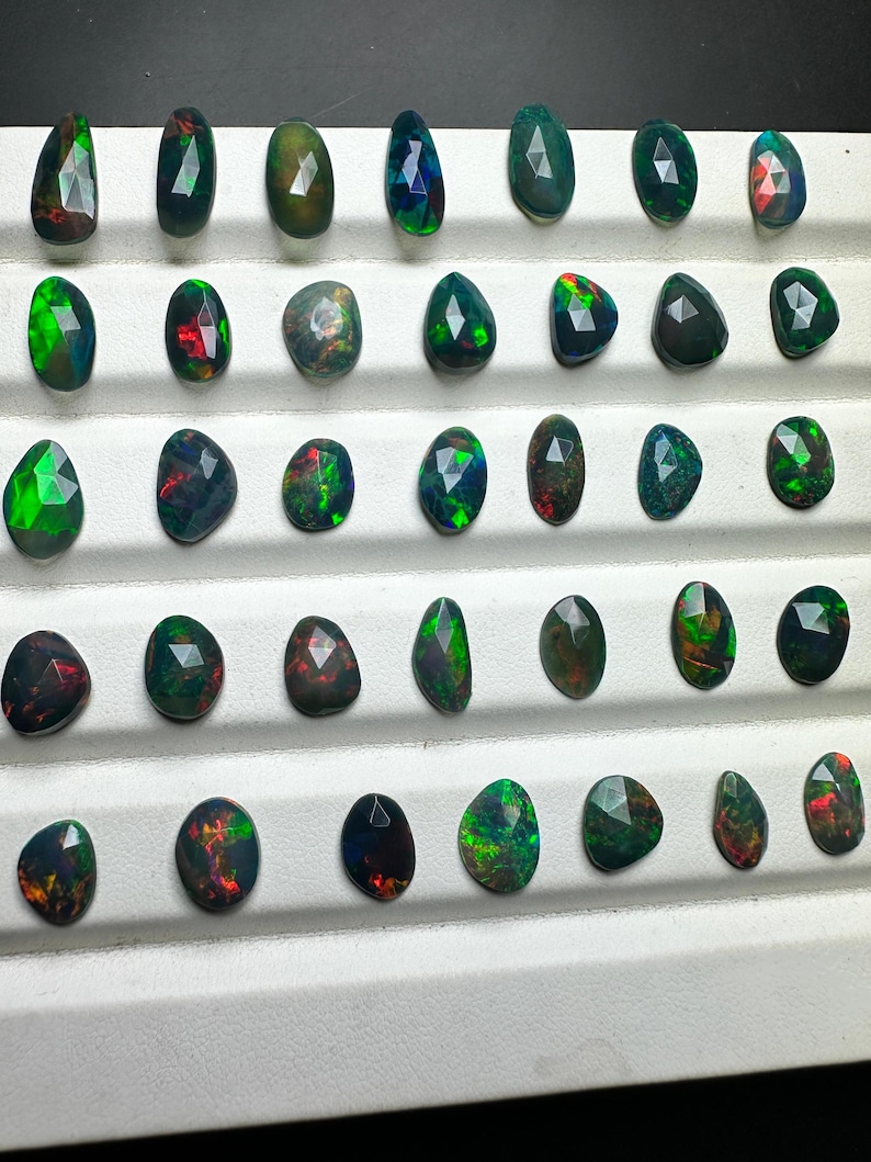 May include: A collection of 36 black opals with green and red flashes. The opals are displayed in rows on a white surface.
