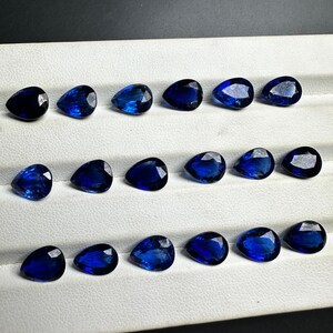 May include: A collection of 18 pear-shaped blue gemstones arranged in three rows on a white surface. The gemstones are all approximately the same size and shape.