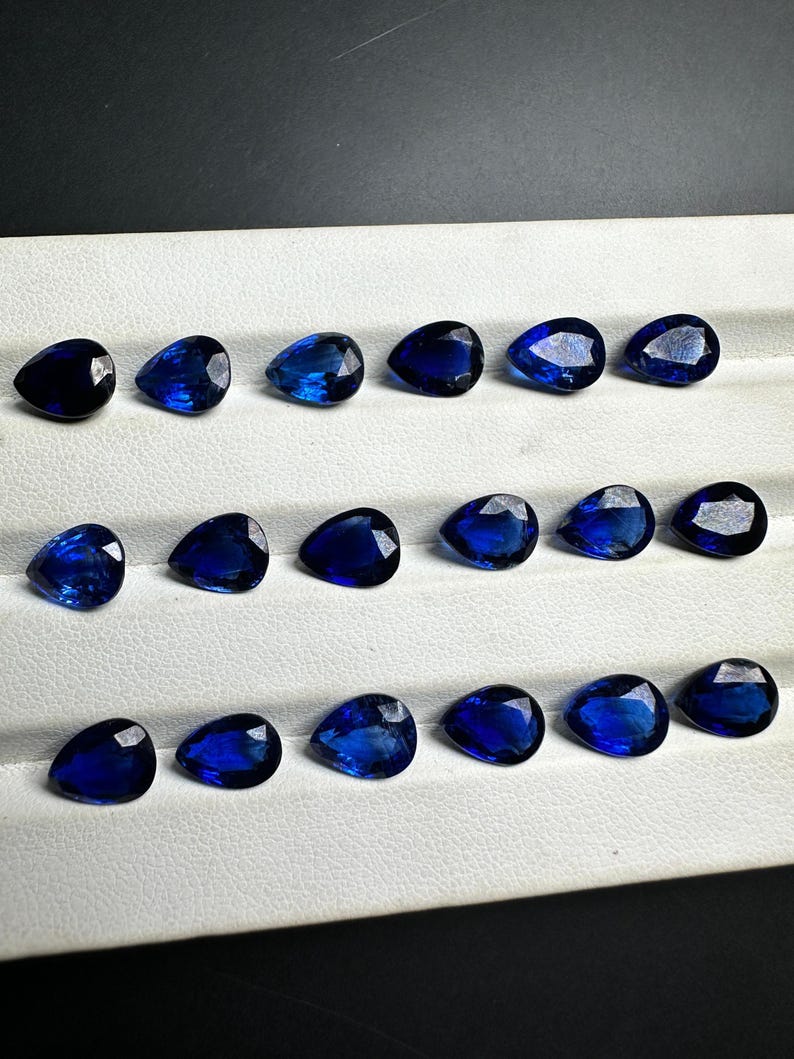 May include: A collection of 18 pear-shaped blue gemstones arranged in three rows on a white surface. The gemstones are all approximately the same size and shape.