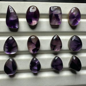 May include: An assortment of amethyst gemstones in various shapes, including oval, teardrop, and rectangular. The stones display a range of purple hues, from deep violet to lighter shades, with some showing internal patterns. The gemstones are arranged on a white surface.