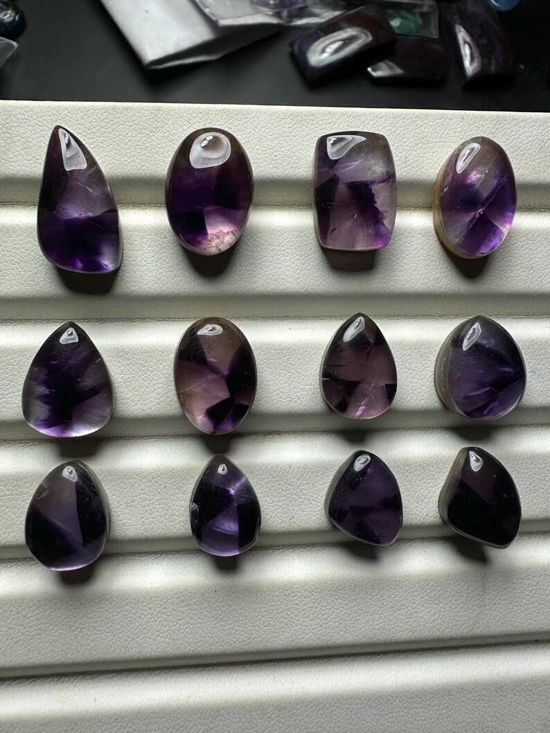 May include: An assortment of amethyst gemstones in various shapes, including oval, teardrop, and rectangular. The stones display a range of purple hues, from deep violet to lighter shades, with some showing internal patterns. The gemstones are arranged on a white surface.