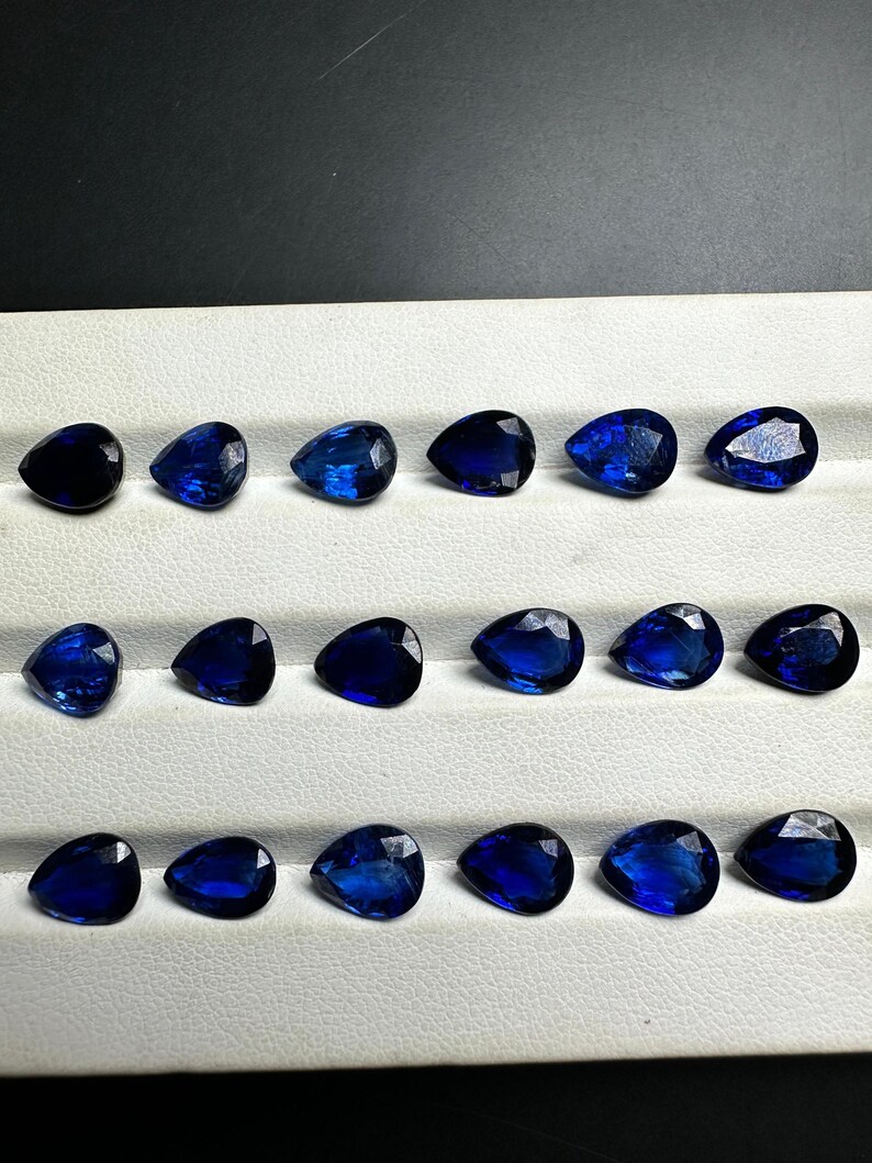 May include: A collection of 15 pear-shaped blue gemstones arranged in three rows of five. The gemstones are displayed on a white surface.