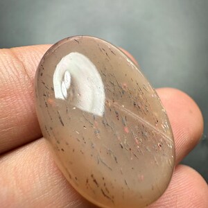 Op de afbeelding: Ovale, doorschijnende edelsteen met een lichtbruine tint. Het heeft interne insluitsels van donkere en rode minerale afzettingen, wat een gespikkelde uitstraling geeft. Het gepolijste oppervlak reflecteert licht en toont natuurlijke patronen.