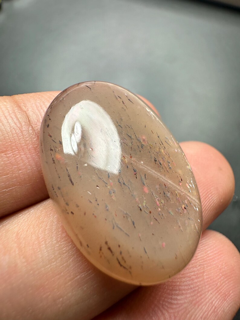 Op de afbeelding: Ovale, doorschijnende edelsteen met een lichtbruine tint. Het heeft interne insluitsels van donkere en rode minerale afzettingen, wat een gespikkelde uitstraling geeft. Het gepolijste oppervlak reflecteert licht en toont natuurlijke patronen.