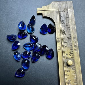 May include: A collection of 14 blue pear-shaped gemstones, measured with a ruler in inches, on a dark gray surface.