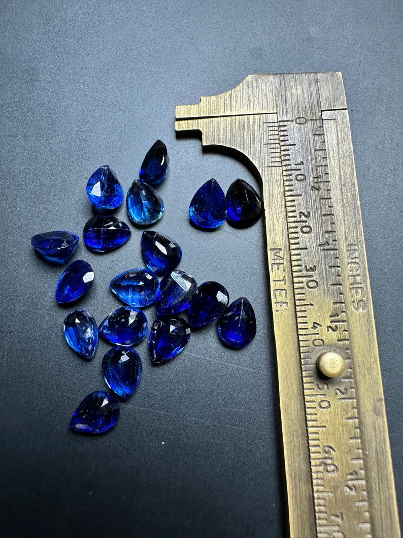 May include: A collection of 14 blue pear-shaped gemstones, measured with a ruler in inches, on a dark gray surface.