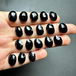 May include: A collection of 15 black faceted oval gemstones. The stones are arranged in rows on a hand.
