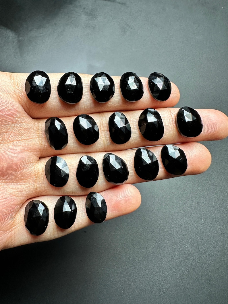 May include: A collection of 15 black faceted oval gemstones. The stones are arranged in rows on a hand.