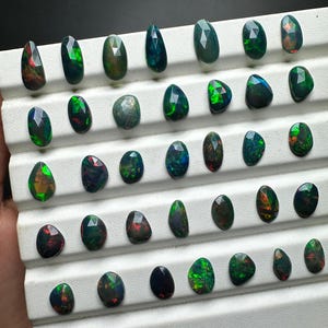 May include: A collection of 40 black opal gemstones, each with a unique green and red iridescent sheen, displayed on a white tray.