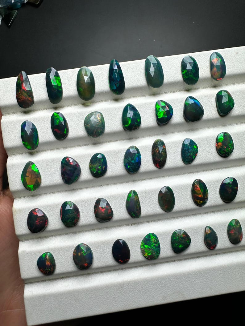 May include: A collection of 40 black opal gemstones, each with a unique green and red iridescent sheen, displayed on a white tray.