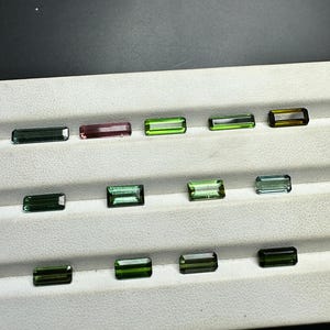 May include: A collection of 14 green, blue, and pink gemstones arranged in rows on a white surface. The gemstones are rectangular in shape and have a smooth, polished finish.