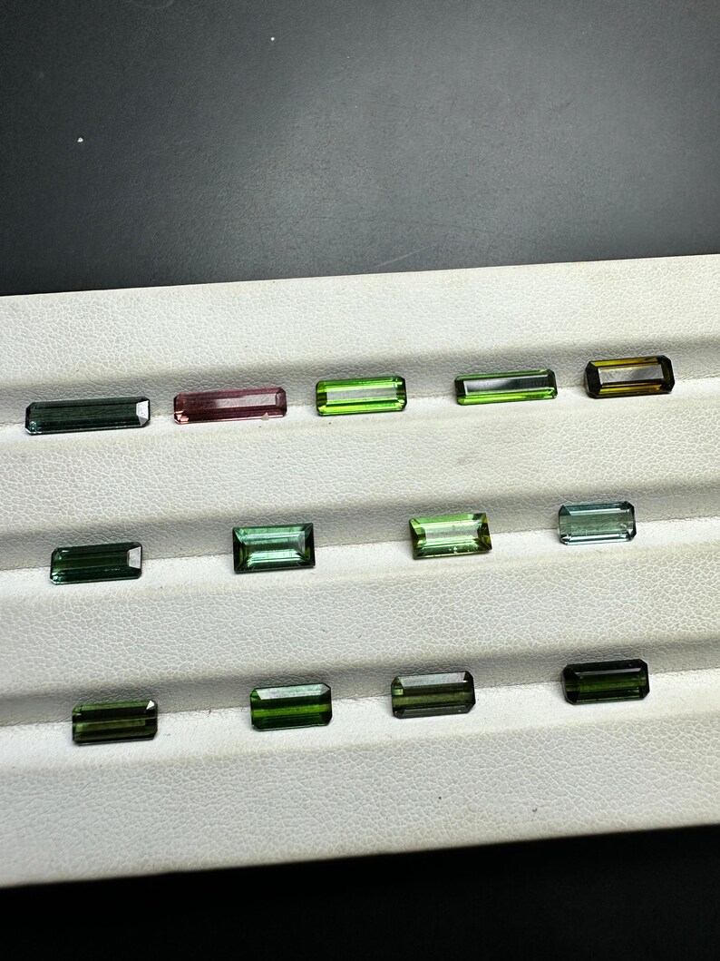 May include: A collection of 14 green, blue, and pink gemstones arranged in rows on a white surface. The gemstones are rectangular in shape and have a smooth, polished finish.