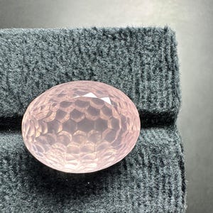 以下が含まれることがあります： ファセット加工が施された、楕円形のローズクォーツの宝石。宝石の表面には、複雑な幾何学模様のファセットがあり、キラキラとした効果を生み出しています。濃いグレーのテクスチャード加工された布の上に置かれています。