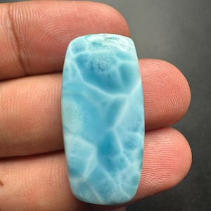 May include: A rectangular cabochon of polished Larimar gemstone featuring a vibrant sky-blue color with intricate white, web-like patterns across its smooth, glossy surface. The stone has rounded corners and a flat, polished face.