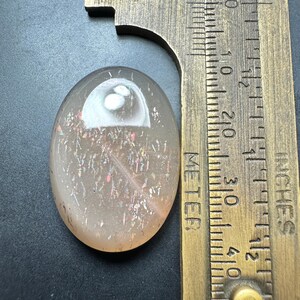 Op de afbeelding: Een ovaalvormige, doorschijnende edelsteen met interne iriserende insluitsels. De steen is lichtoranje met roze en gouden flitsen. Een messing meetinstrument ligt naast de steen en geeft centimeters aan. De steen is ongeveer 3,8 cm lang.