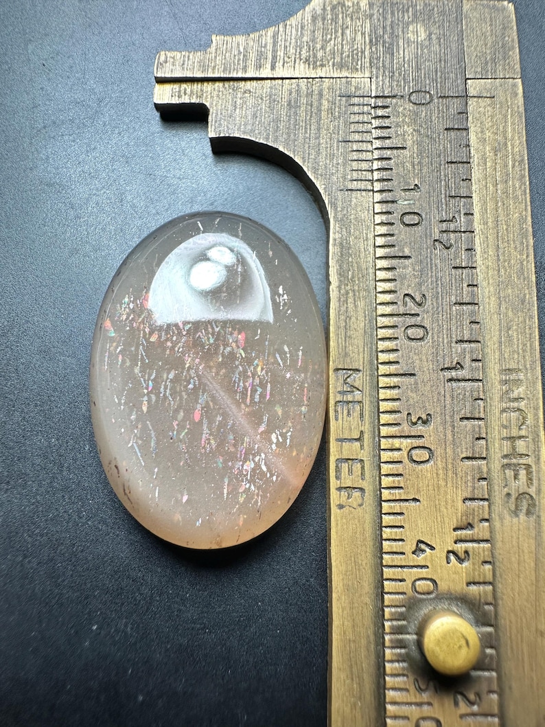Op de afbeelding: Een ovaalvormige, doorschijnende edelsteen met interne iriserende insluitsels. De steen is lichtoranje met roze en gouden flitsen. Een messing meetinstrument ligt naast de steen en geeft centimeters aan. De steen is ongeveer 3,8 cm lang.