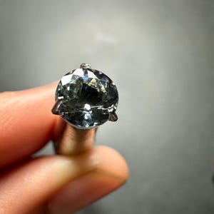 May include: A round, faceted gemstone with a dark blue-green colour, set in a silver prong setting.