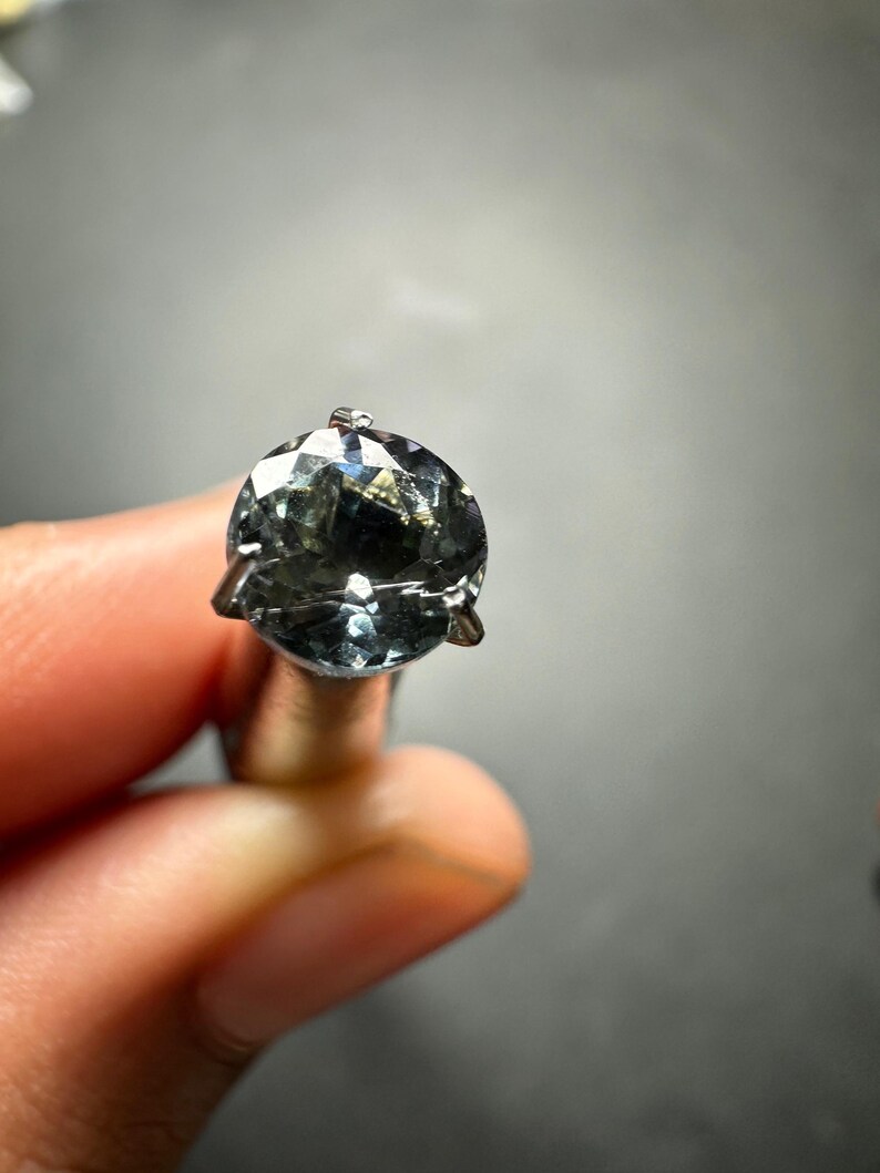 May include: A round, faceted gemstone with a dark blue-green colour, set in a silver prong setting.