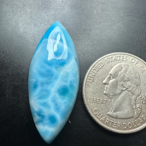 May include: A marquise-shaped, polished blue and white Larimar gemstone cabochon with a smooth, glossy surface, positioned next to a United States quarter dollar coin for scale to show its relative size.