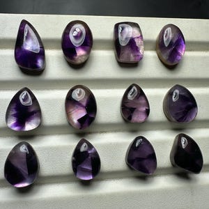 May include: An assortment of polished amethyst gemstones in various shapes, including teardrop, oval, and rectangular. The stones display a range of purple hues with translucent and opaque sections. The gemstones are arranged on a white tray.