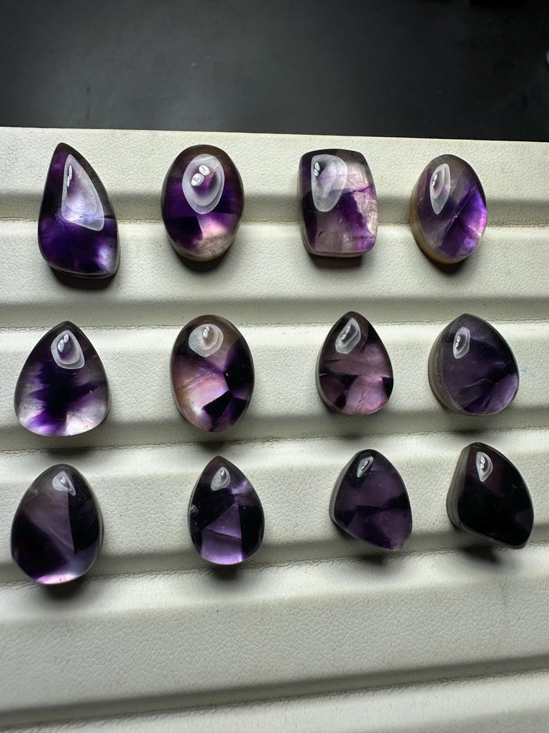 May include: An assortment of polished amethyst gemstones in various shapes, including teardrop, oval, and rectangular. The stones display a range of purple hues with translucent and opaque sections. The gemstones are arranged on a white tray.