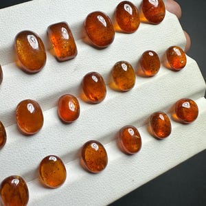 May include: A collection of 15 oval and rectangular shaped gemstones in various shades of brown. The gemstones are displayed on a white surface.
